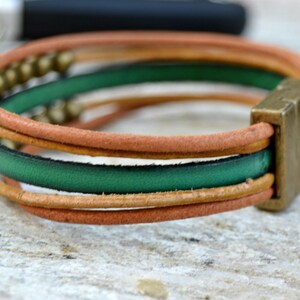 May include: Leather bracelet with a square metal clasp. The bracelet features three bands: tan, green, and tan. Small gold-colored beads are visible on the top band. The bracelet is on a light-colored surface.