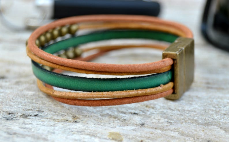 May include: Leather bracelet with a square metal clasp. The bracelet features three bands: tan, green, and tan. Small gold-colored beads are visible on the top band. The bracelet is on a light-colored surface.