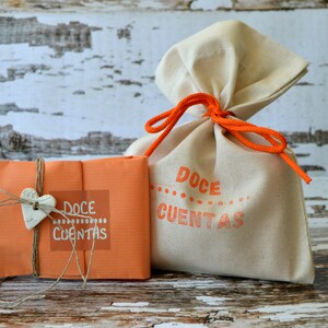 May include: Two gift bags. One is a beige drawstring bag tied with orange cord. The other is an orange package tied with twine and a heart-shaped charm. Both have the words "Doce Contas" printed on them in orange.
