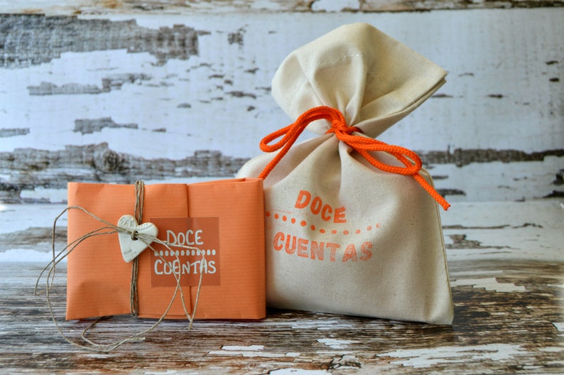 May include: Two gift bags. One is a beige drawstring bag tied with orange cord. The other is an orange package tied with twine and a heart-shaped charm. Both have the words "Doce Contas" printed on them in orange.