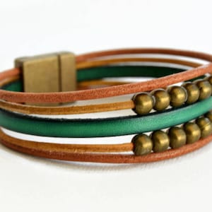 May include: A multi-strand bracelet featuring brown, green, and gold leather cords. The bracelet has a magnetic clasp and is adorned with gold-colored beads. The design is a mix of textures and colors.