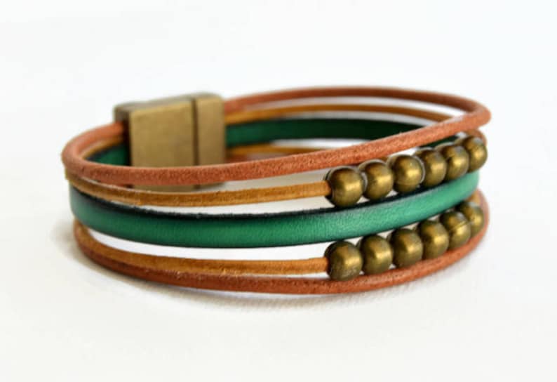 May include: A multi-strand bracelet featuring brown, green, and gold leather cords. The bracelet has a magnetic clasp and is adorned with gold-colored beads. The design is a mix of textures and colors.