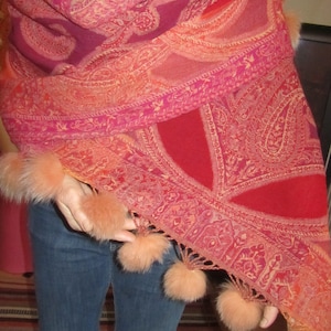 May include: A red, pink, and orange paisley patterned shawl with a fringe border and fluffy pom-poms. The shawl is draped over a person's shoulders and is worn with blue jeans.