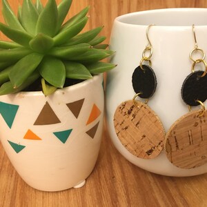 May include: A pair of dangle earrings with a black leather circle and a cork circle. The earrings have gold-colored metal hoops and chains.