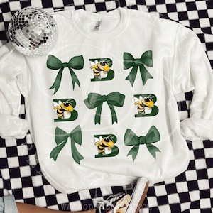 Puede incluir: Sudadera blanca con un gráfico de abeja verde y amarilla y lazos verdes. El gráfico de la abeja es una letra B con una abeja en ella. Los lazos están en un patrón de cuadrícula en la sudadera.