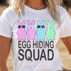 Puede incluir: Camiseta blanca con un gráfico de cuatro conejitos de Pascua de dibujos animados con gafas de sol. El texto "EGG HIDING SQUAD" está impreso debajo de los conejos. La persona lleva un sombrero de vaquero.