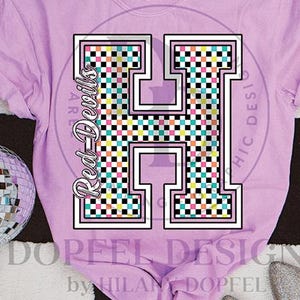 May include: A light purple t-shirt featuring a large, white-outlined letter "H" filled with a colorful checkered pattern. The text "Red Devils" is printed vertically on the left side of the "H". A disco ball is visible on the left.
