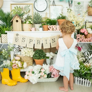 May include: A young girl in a white dress with a blue bow stands in front of a white shelf with a spring banner that reads "SPRING". The shelf is decorated with flowers, plants, and other spring-themed items.
