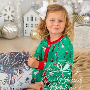 May include: A young girl wearing a green and red Christmas pajamas with a Nutcracker and ballerina print is sitting on a tan blanket and reading a book. The book has a colorful illustration of a winter scene with trees and snow.