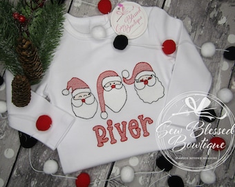 Puede incluir: Mono blanco de manga larga con tres caras de Papá Noel bordadas y el nombre "River" en bordado rojo.
