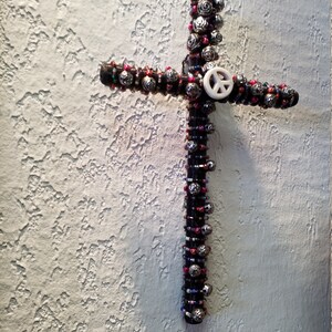 May include: A beaded cross with a peace sign in the centre. The cross is made of black wire and is decorated with silver, pink, and blue beads.