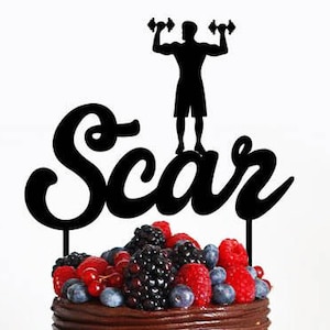 May include: Black silhouette of a person lifting weights with the name "Scar" written in a cursive font. The cake topper is on top of a chocolate cake with berries.