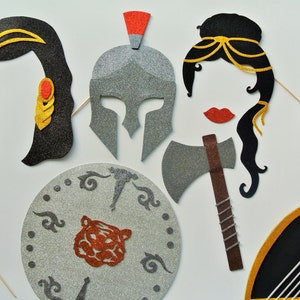 Puede incluir: Un conjunto de seis accesorios de cabina de fotos de fieltro con temática de mitología griega. Los accesorios incluyen un casco de purpurina plateada, un hacha de purpurina plateada, un arpa marrón y dorada, un escudo de purpurina plateada con un diseño de tigre y dos piezas de cabello negras con detalles dorados.