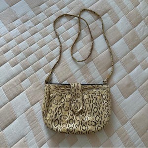 May include: A small crossbody bag featuring a tan and black snakeskin pattern. The bag has a rounded bottom, a thin matching shoulder strap, and a central vertical tab closure with a metallic gold-tone accent at the bottom of the tab.