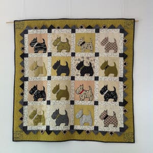 Op de afbeelding: Een handgemaakte quilt met een herhalend patroon van terri&euml;r honden applicaties in verschillende kleuren en patronen. De quilt heeft een geelgroene rand en is opgehangen aan een houten deuvel. De honden staan in vierkanten.
