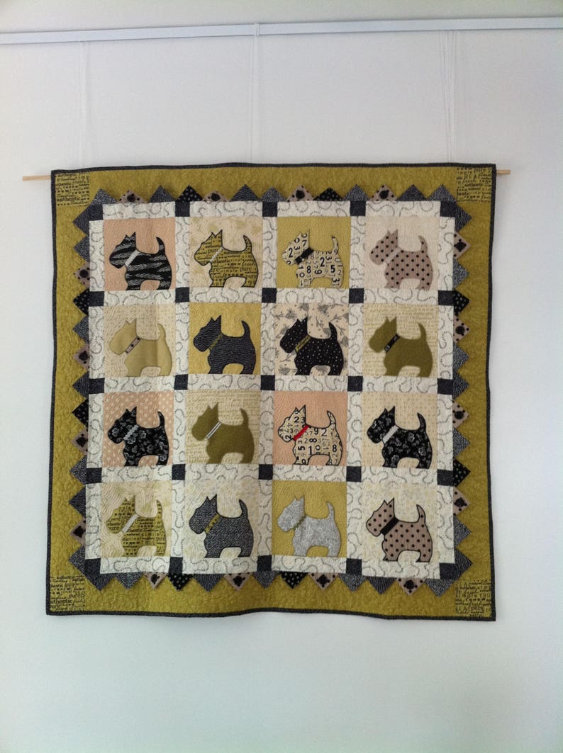 Op de afbeelding: Een handgemaakte quilt met een herhalend patroon van terri&euml;r honden applicaties in verschillende kleuren en patronen. De quilt heeft een geelgroene rand en is opgehangen aan een houten deuvel. De honden staan in vierkanten.