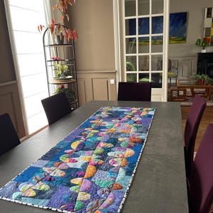 Op de afbeelding: Een kleurrijke patchwork tafelrunner met een geometrisch ontwerp in tinten blauw, paars, groen en geel. De runner ligt op een grijze tafel met stoelen.