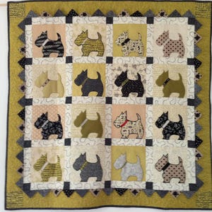 Op de afbeelding: Een handgemaakte quilt met een herhalend patroon van terriër honden applicaties. De honden zijn in verschillende kleuren en patronen, tegen een patchwork achtergrond van neutrale tinten. De quilt heeft een decoratieve rand in groen- en grijstinten.