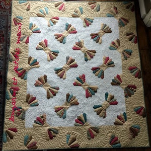 Könnte beinhalten: Eine weiße Steppdecke mit einem beigefarbenen Rand und einem sich wiederholenden Muster aus bunten fächerförmigen Designs. Die Steppdecke ist mit einem wirbelnden Muster gesteppt. Der Text "long point et petits mains" ist in Pink auf der linken Seite der Steppdecke geschrieben.