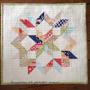 Può includere: Una trapunta bianca con un motivo a stella a raggiera in tonalità di rosso, blu, verde e beige. La trapunta è bordata con un bordo verde e rosa. Il testo "longarm et petites mains" è scritto in inchiostro blu nell'angolo in basso a destra.
