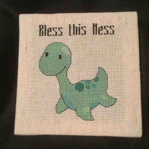 May include: A cross-stitch embroidery on canvas with a green dinosaur and the text "Bless this mess".