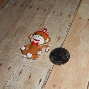 May include: A brown monkey figurine with a red and white striped hat, a red bow tie, and white socks. The figurine is sitting with its legs crossed and its arms outstretched. The figurine is about the size of a US quarter.