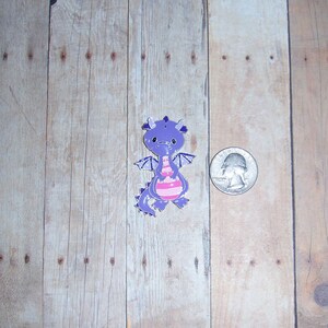 May include: Purple and pink cartoon dragon charm with a white background. The charm is about the size of a US quarter.