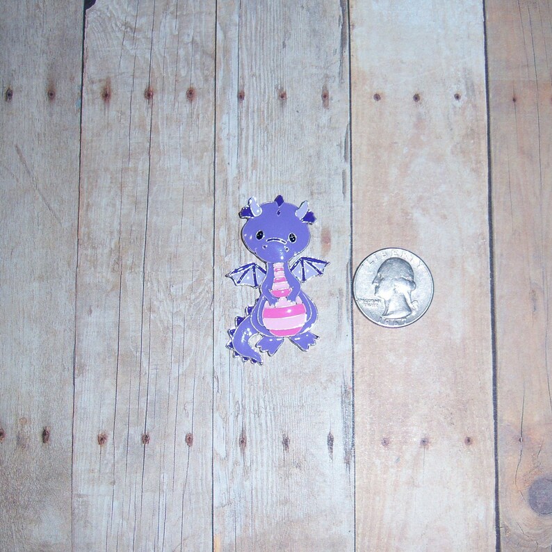 May include: Purple and pink cartoon dragon charm with a white background. The charm is about the size of a US quarter.