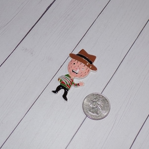 May include: A red, green, and white striped shirt with a brown hat and a smiling face. The figure is holding a knife in its right hand. The figure is made of metal and is a pin.