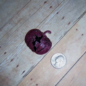 May include: A purple pumpkin with a black spider web and a black silhouette of a bat on the front. The pumpkin is about the size of a quarter.