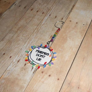 May include: A white Christmas ornament with a circle of colorful lights around the edge. The ornament says "Friends Don't Lie" in black text. The ornament has a silver hanger with a rainbow of beads.