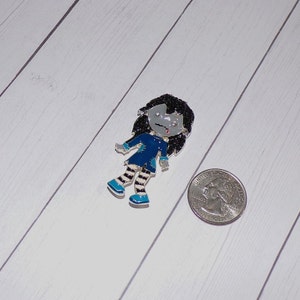 May include: A silver charm of a cartoon girl with black hair, a blue dress, and white and black striped stockings. The charm is about the size of a quarter.