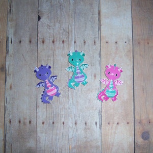 May include: Three colorful dragon figurines with pink, green, and purple paint. The dragons have wings and are facing forward. The dragons are on a wooden background.