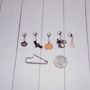 May include: Set of seven Halloween-themed stitch markers with charms including a bat, pumpkin, ghost, cat, and lips. The markers are silver with a spring-loaded clasp. A silver safety pin is also included.