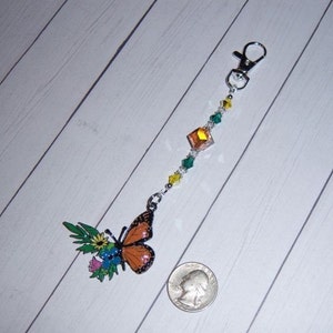 May include: A silver key chain with a butterfly charm. The butterfly is orange, black, and white with a floral design. The chain is decorated with green, yellow, and clear crystals.
