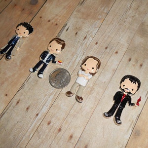 May include: Four cartoon character pins. Each pin features a different character in a suit or casual attire. The pins are colorful with black, white, red, and silver accents. The pins are on a light brown wooden surface.