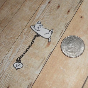 May include: A white enamel pin of a sleeping cat with a silver chain attached to a white enamel charm with the letter Z. The pin is about the size of a US quarter.