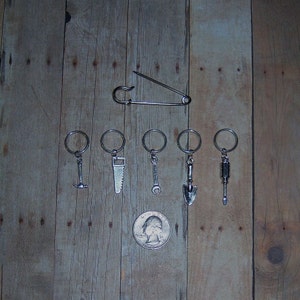 May include: A set of six silver keychains featuring miniature tools, including a hammer, saw, wrench, shovel, and screwdriver. The keychains are displayed on a wooden background with a safety pin.