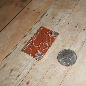 May include: A small, brown, metal book charm with silver accents and a decorative design. The charm is about the size of a US quarter.