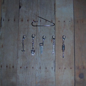 May include: A set of six silver charm pendants featuring a variety of tools, including a shovel, saw, wrench, and screwdriver. The charms are attached to silver jump rings and are ready to be added to a keychain or bracelet.