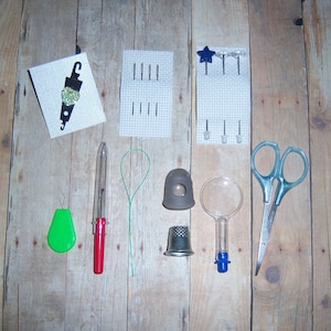 May include: A collection of needlework tools and supplies, including a needle threader, a thimble, a magnifying glass, a pair of scissors, a needle minder, and a small spool of thread.