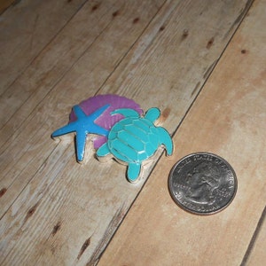 May include: A silver charm with a blue starfish, a purple seashell, and a turquoise sea turtle. The charm is about the size of a US quarter.