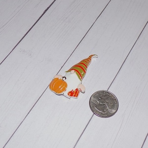 May include: A small, white gnome figurine with a green and red striped hat, holding a small orange pumpkin. The gnome is about the size of a US quarter.