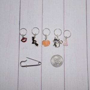 May include: Set of six Halloween-themed stitch markers with charms including a vampire mouth, bat, pumpkin, cat, and ghost. The markers are silver with a silver safety pin.
