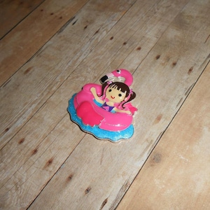 May include: A pink flamingo pool float with a girl in a pink and purple swimsuit sitting inside. The float is on a blue glitter background.