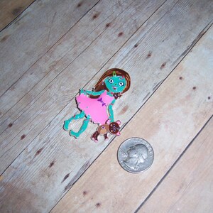 May include: A green zombie girl doll with brown hair wearing a pink dress and holding a brown teddy bear.