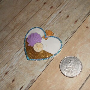 May include: A heart-shaped pin with a white background and a gold glitter design. The pin features a purple seashell, a yellow starfish, and a blue glitter border.
