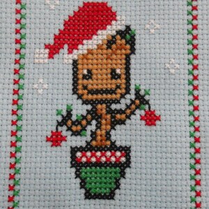 Könnte beinhalten: Kreuzstich eines Charakters mit Weihnachtsmütze, der Beeren hält und in einem grünen Topf steht. Der Charakter ist braun mit schwarzer Umrandung. Der Hintergrund ist hellblau, mit einem rot-grünen Rand.