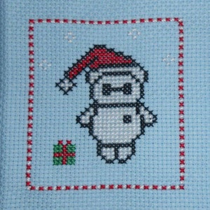 May include: A cross-stitch pattern of a robot wearing a red Santa hat and holding a small gift box. The robot is white with black eyes and a black mouth. The background is light blue.