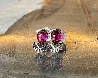 May include: A pair of silver stud earrings featuring teardrop-shaped, vibrant pink gemstones. The earrings have a delicate, floral-inspired design at the base, and are set against a reflective surface.